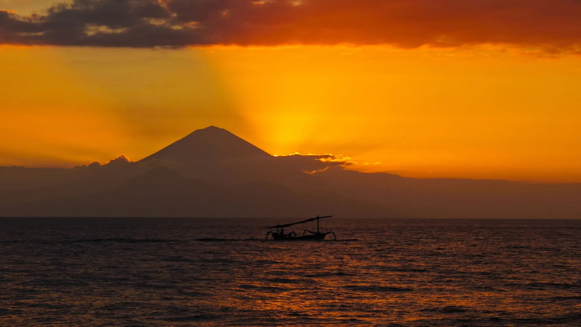 A jukung outrigger boat on the water at sunset below Mount Agung